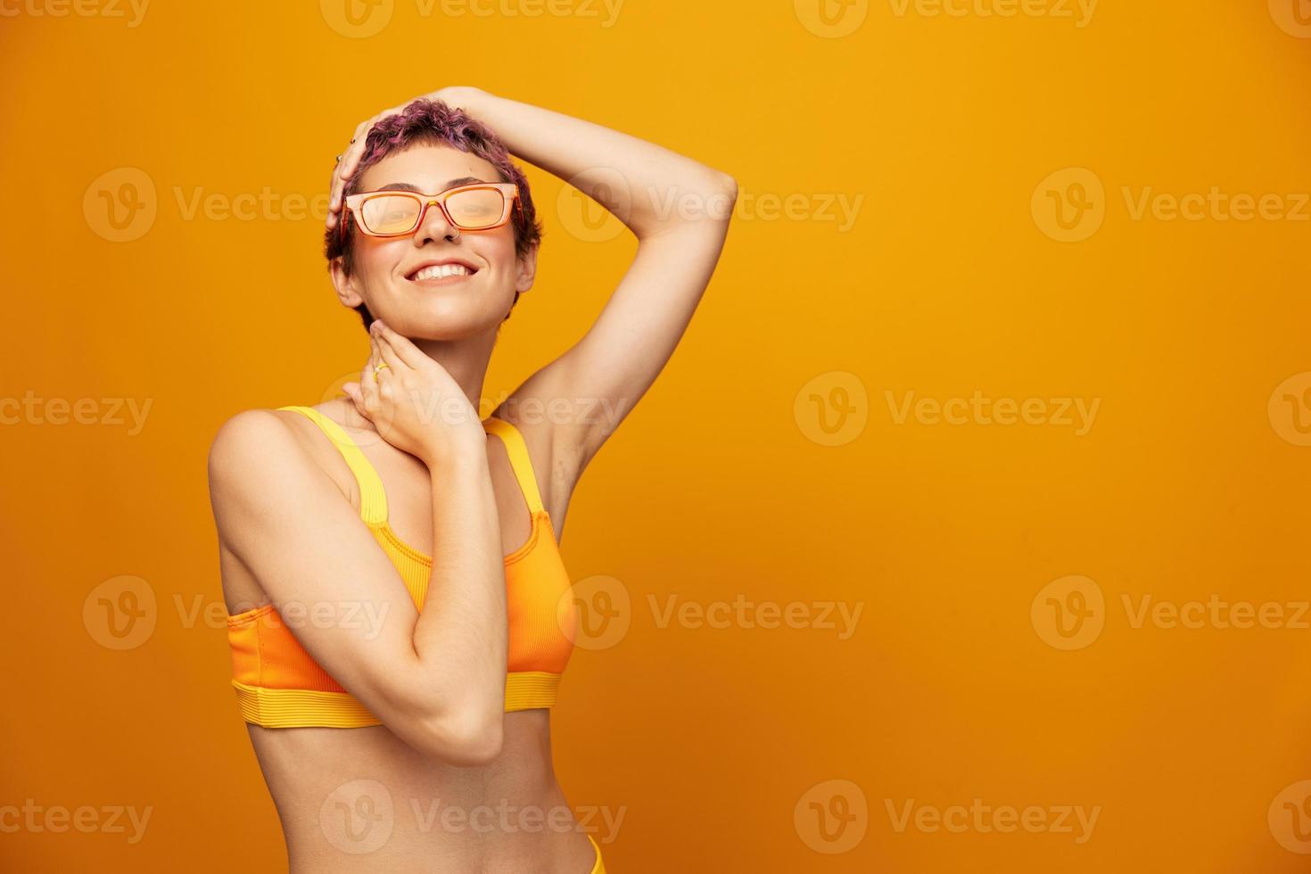 jovem mulher dentro uma brilhante amarelo ginástica fato de treino com a aberto barriga e oculos de sol em a laranja fundo sorridente foto