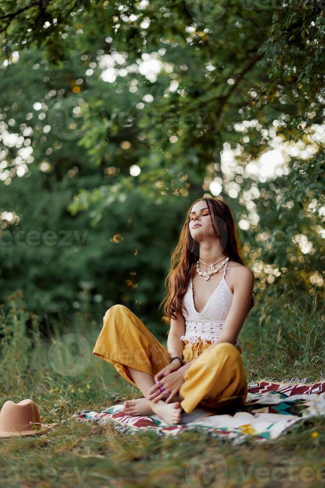 uma jovem hippie mulher medita dentro natureza dentro a parque, sentado dentro uma lótus posição em dela colorida xadrez e desfrutando harmonia com a mundo dentro roupas ecológicas dentro a outono foto
