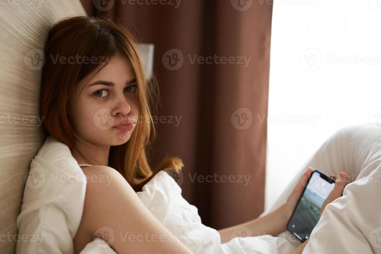 emocional mulher mentiras dentro cama debaixo a cobre com Móvel telefone toque tela interior foto