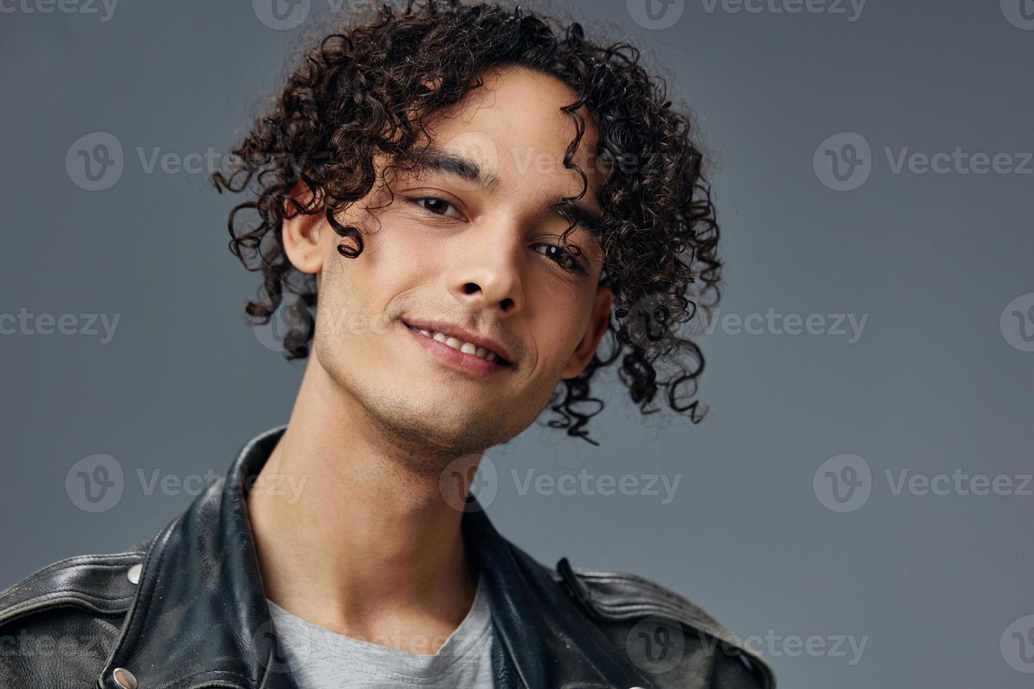 Tiros na Cabeça retrato do à moda bronzeada encaracolado homem couro Jaqueta posando isolado em sobre cinzento estúdio fundo. legal moda oferecer. enorme sazonal venda Novo coleção conceito. cópia de espaço para de Anúncios foto