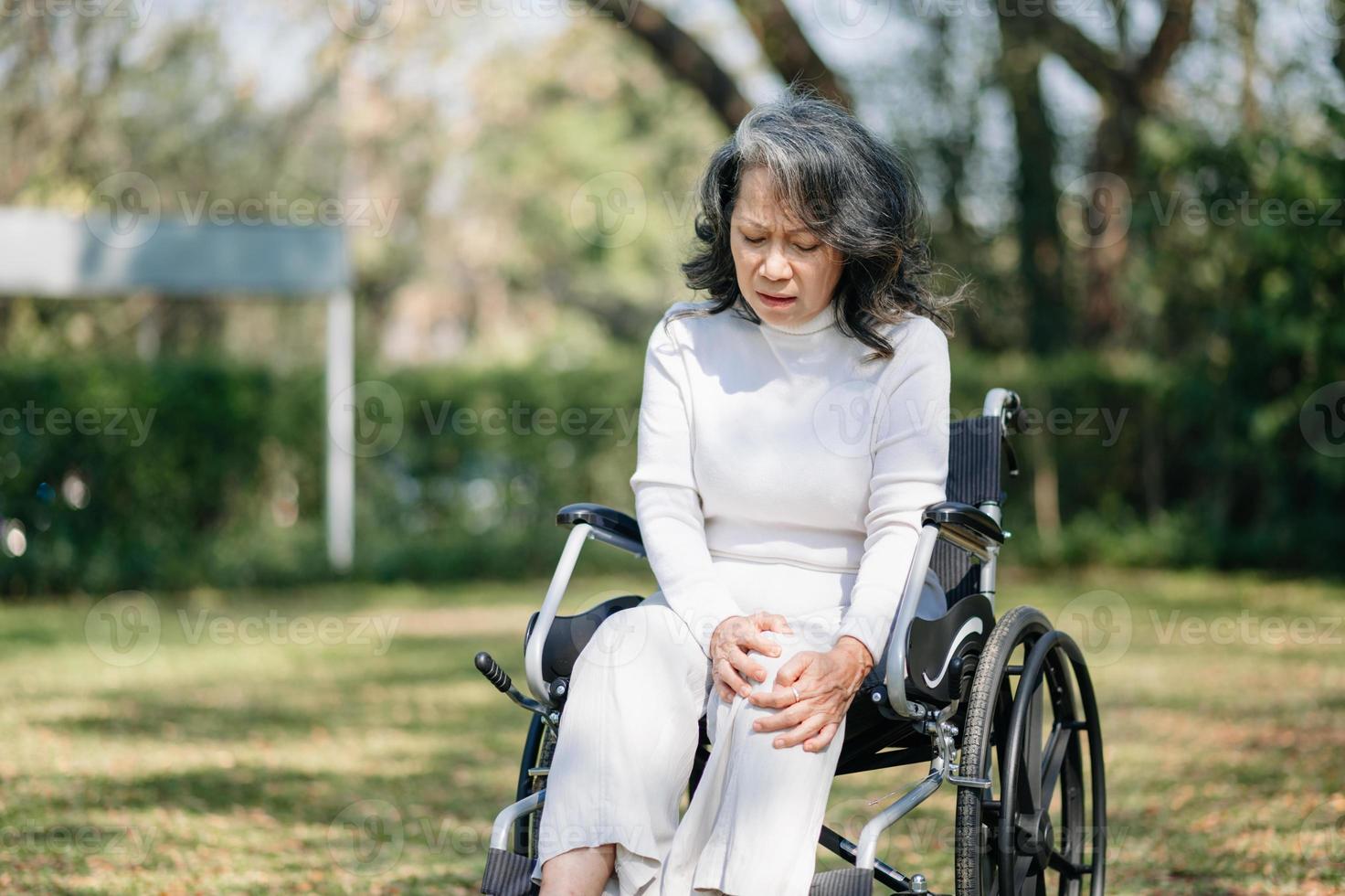 ásia velho mulher sentado em uma cadeira de rodas ao ar livre dentro a parque ter dor dentro a braços, pulsos e corpo dentro Sol luz foto