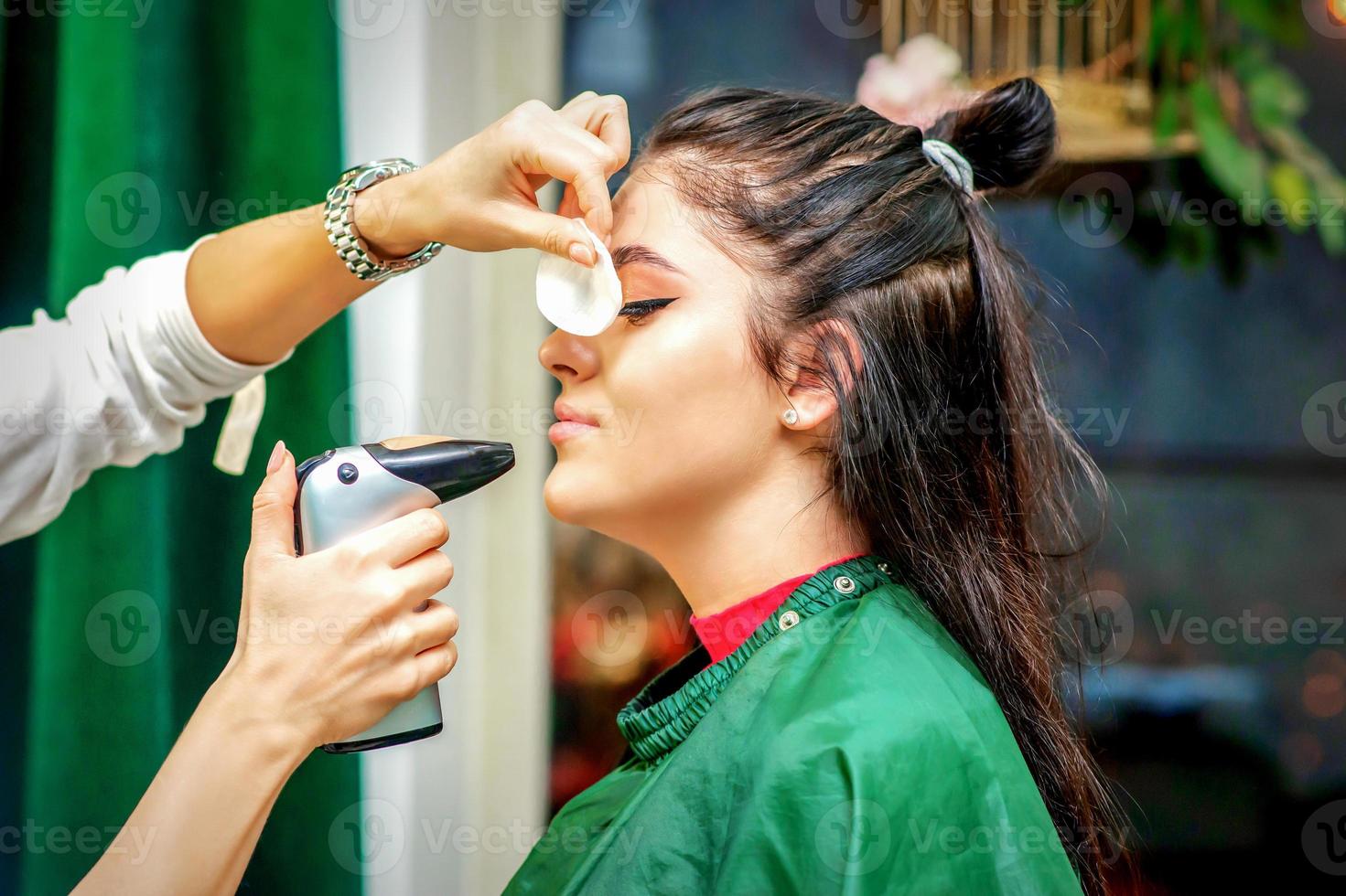 lado Visão do uma Maquiagem artista usando aerógrafo fazer a aerógrafo Maquiagem Fundação em uma fêmea face dentro uma beleza salão. foto