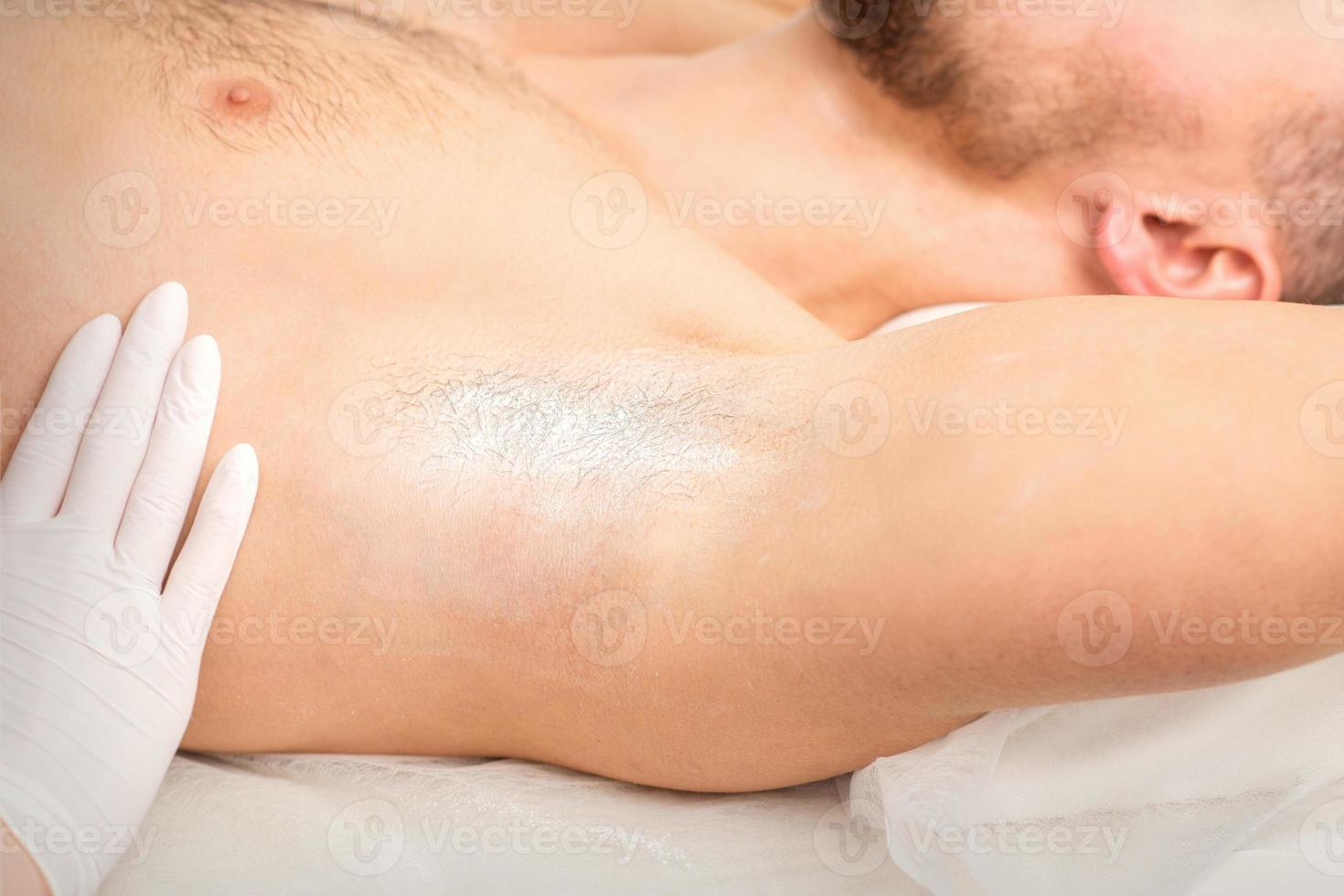 jovem caucasiano homem recebendo cabelo remoção a partir de dele axila dentro uma beleza salão, depilação masculino axila. foto