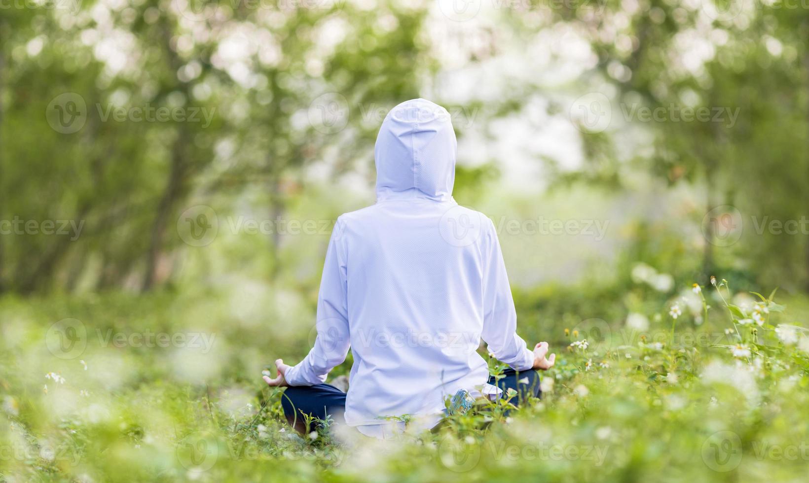 traseiro Visão do mulher dentro moletom com capuz é relaxantemente praticando meditação ioga dentro floresta cheio do margarida flor dentro verão para atingir felicidade a partir de interior Paz sabedoria com manhã luz para saudável mente e alma foto