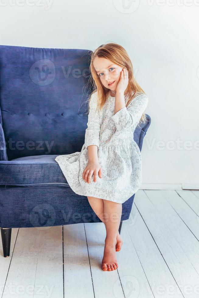 doce pequeno menina dentro branco vestir sentado em moderno acolhedor azul cadeira relaxante dentro branco brilhante vivo quarto às casa dentro de casa. infância escolares juventude relaxar conceito. foto