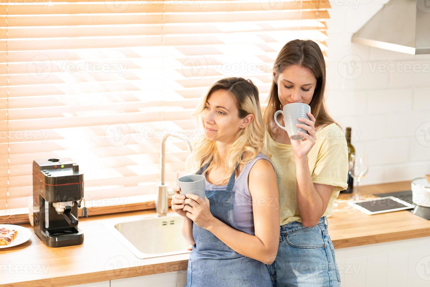 feliz lésbica casal segurando copos do café dentro cozinha. casal do lésbica meninas apreciar café às casa levando sobre algo. dois jovem adulto lindo mulheres beber chá dentro moderno cozinha. foto