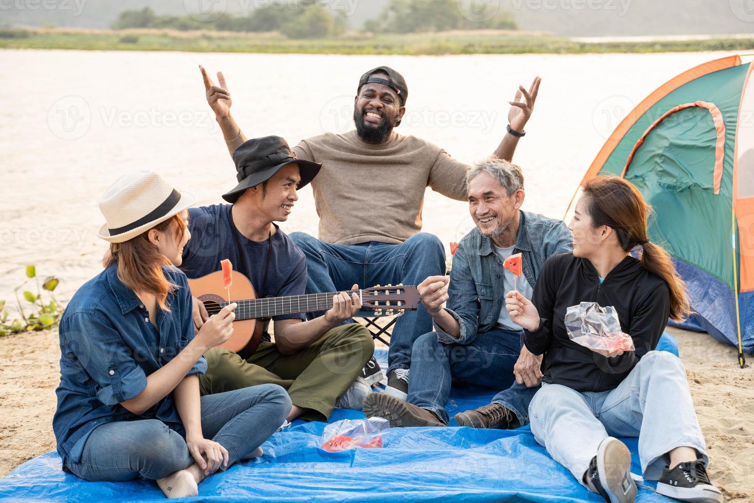 feliz amigos sentado em a de praia cantando e jogando guitarra durante a pôr do sol foto