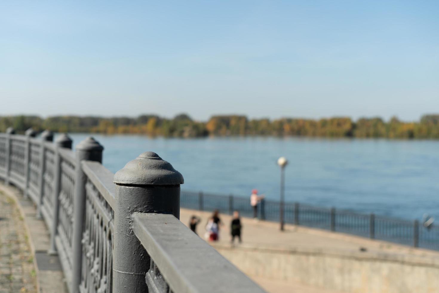 paisagem urbana de uma cerca de metal ao lado de um corpo d'água com pessoas desfocadas ao fundo e um céu azul claro em irkutsk, na Rússia foto