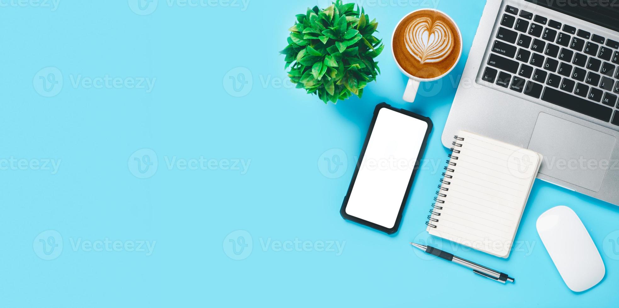 mesa de escritório com computador portátil, telefone inteligente de tela em branco, xícara de café, caneta, caderno sobre fundo azul, vista superior com espaço de cópia, mock up. foto