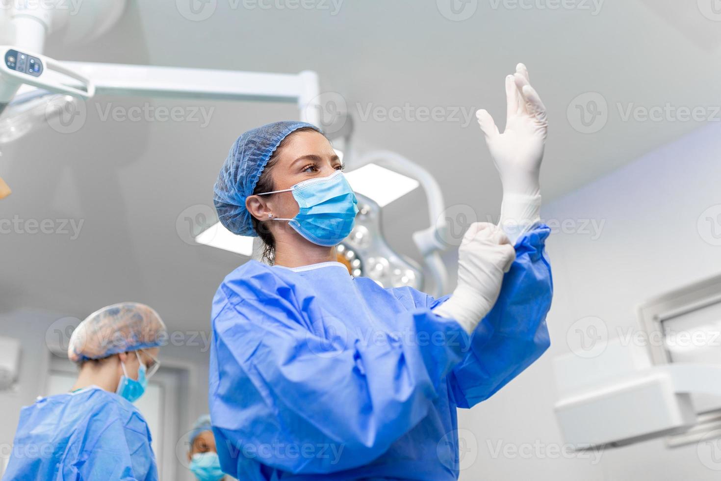 retrato da bela médica cirurgiã calçar luvas médicas em pé na sala de operação. cirurgião na sala de cirurgia moderna foto