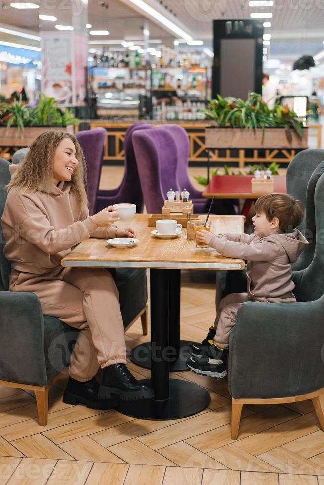 jovem encaracolado cabelos mãe e dela pequeno filho ter café da manhã ou almoço dentro uma cafeteria, sentado às uma mesa foto