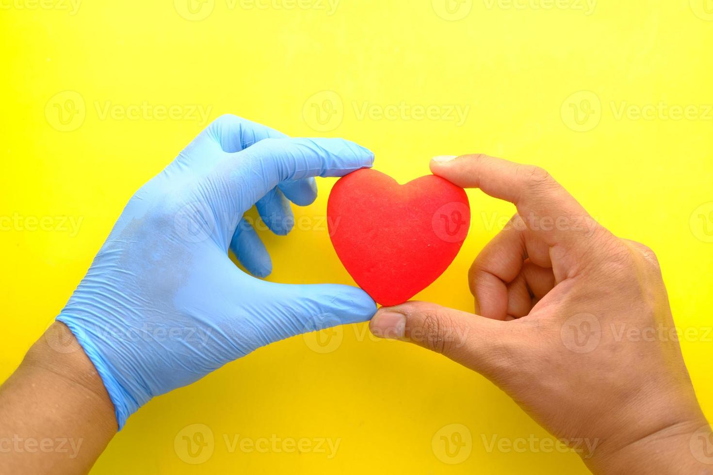 mão do homem em luvas de proteção segurando um coração vermelho sobre fundo amarelo foto