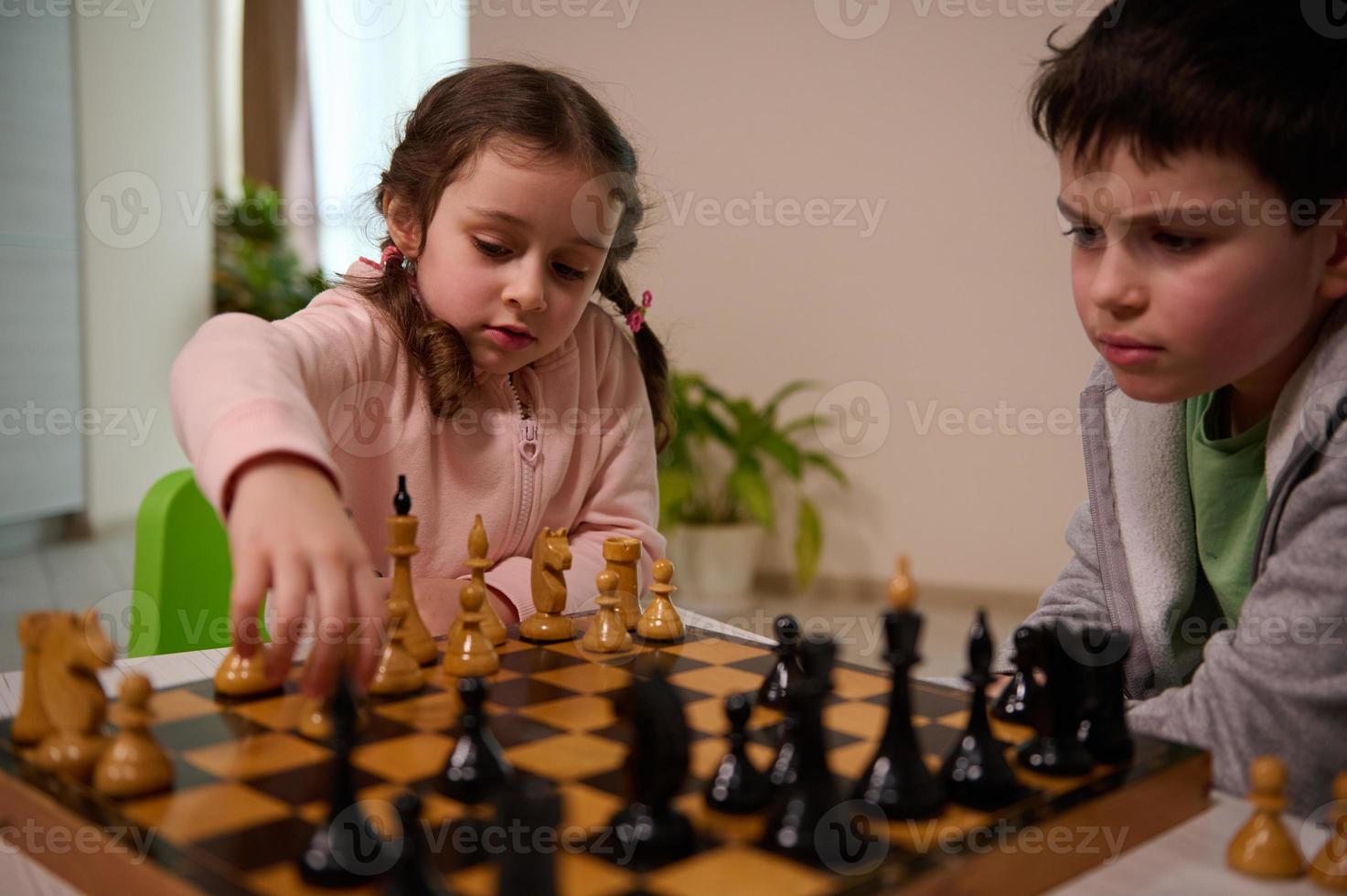 fofa pequeno menina com sério Veja sentado às a mesa e tocam xadrez com dela irmão, colheita acima uma xadrez peça e fazer dele mover. cedo desenvolvimento, casa educacional jogos para crianças foto