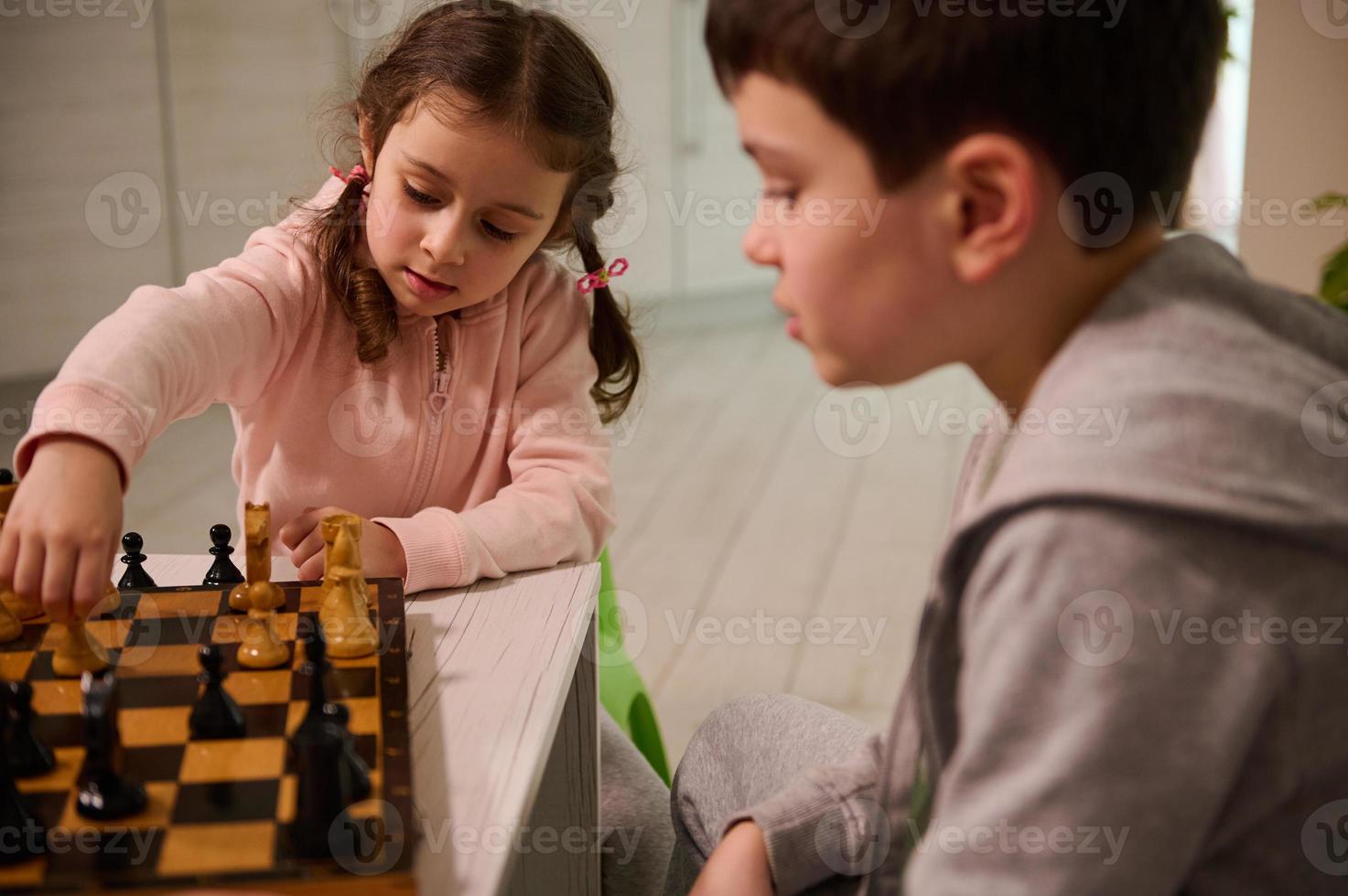 retrato do uma concentrado 4 anos velho pequeno menina colheita acima uma xadrez peça enquanto fazer mover em xadrez quadro, jogando com dela irmão quem ensina dela a jogos foto