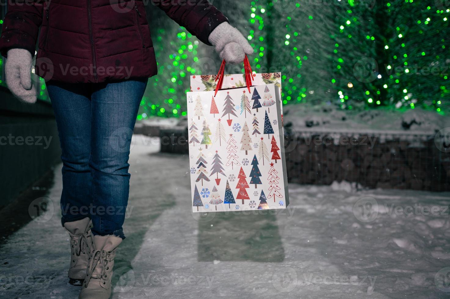 compras bolsas com Natal padronizar dentro mãos do uma mulher, caminhando baixa a neve coberto cidade rua às inverno noite foto