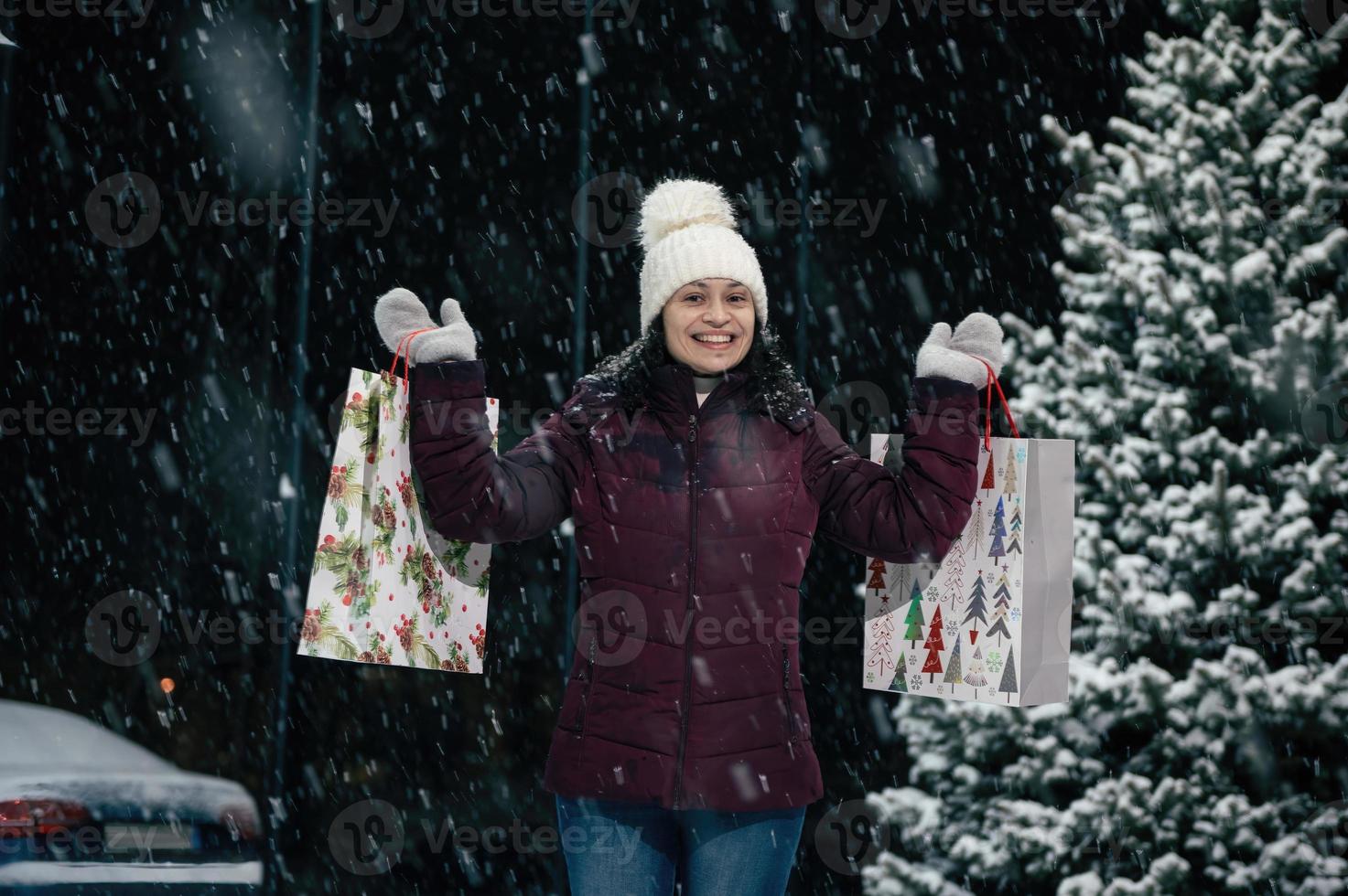 lindo m mulher dentro caloroso roupas, lã chapéu e luvas, com compras bolsas dentro dela mãos, em uma Nevado inverno tarde foto