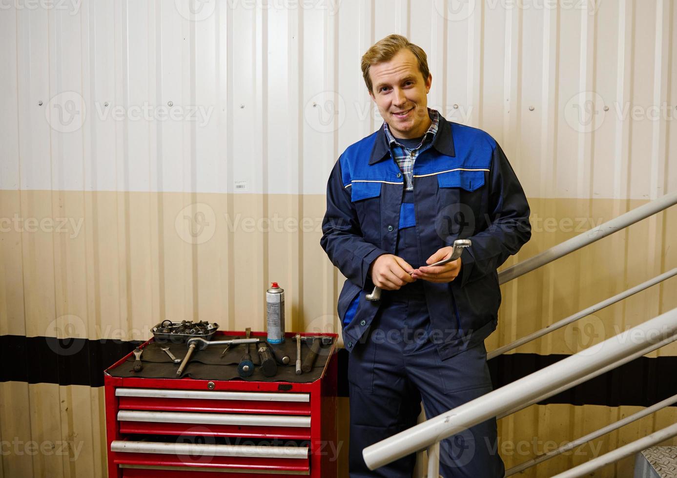 retrato do uma bonito caucasiano homem, garagem mecânico, técnico segurando chave inglesa e olhando às Câmera em pé perto uma caixa com trabalhos Ferramentas para inspecionar e reparar automóveis e carro serviço estação foto