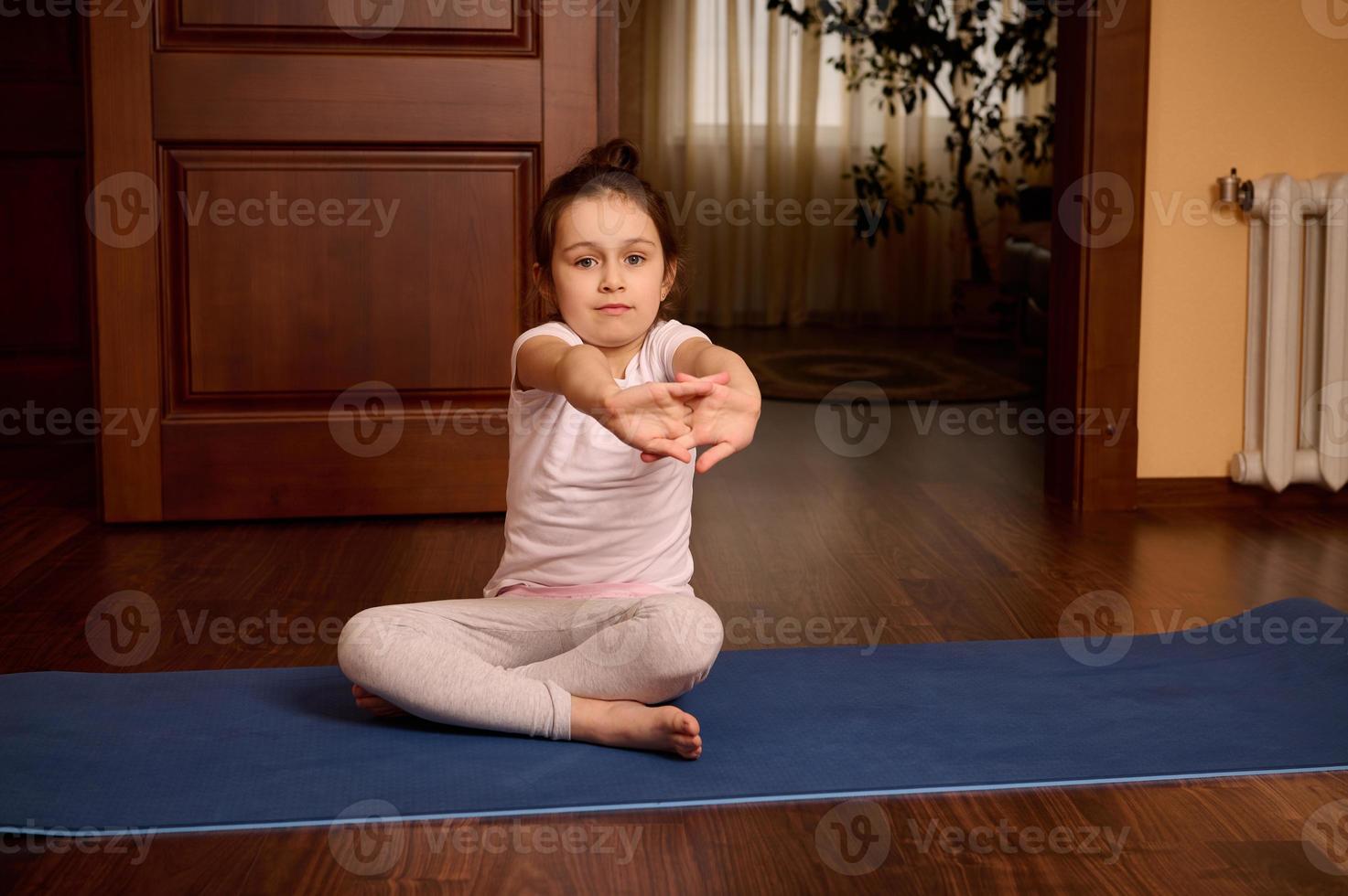 adorável pequeno criança menina sentado em ginástica esteira, alongamento dela braços enquanto exercício, praticando manhã ioga às casa foto