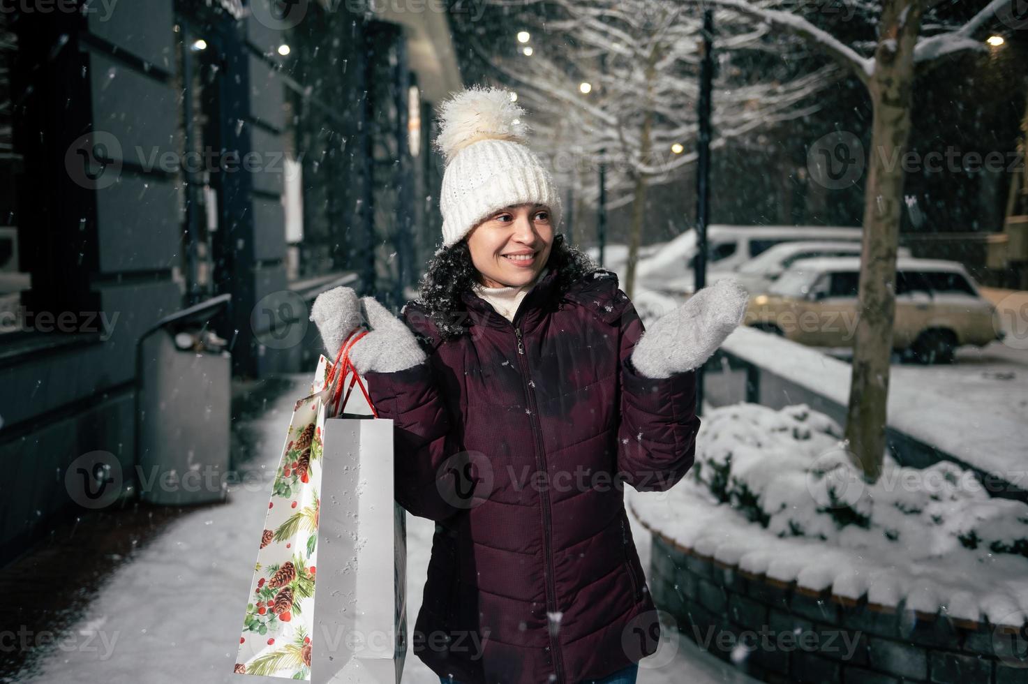 atraente mulher com compras bolsas, pega flocos de neve enquanto anda em ao longo uma Nevado rua iluminado de feriado guirlandas foto