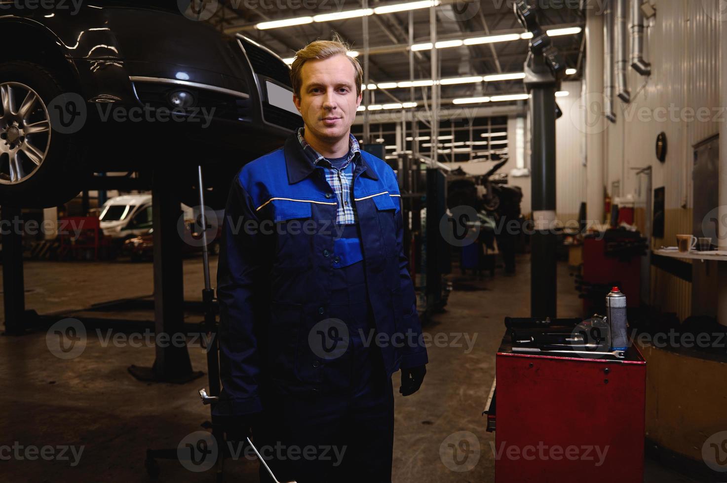 uma técnico, auto mecânico, carro engenheiro em pé contra a fundo do uma levantado automóvel com chave inglesa dentro dele mão, trabalhando dentro reparar fazer compras ou carro serviço garagem foto