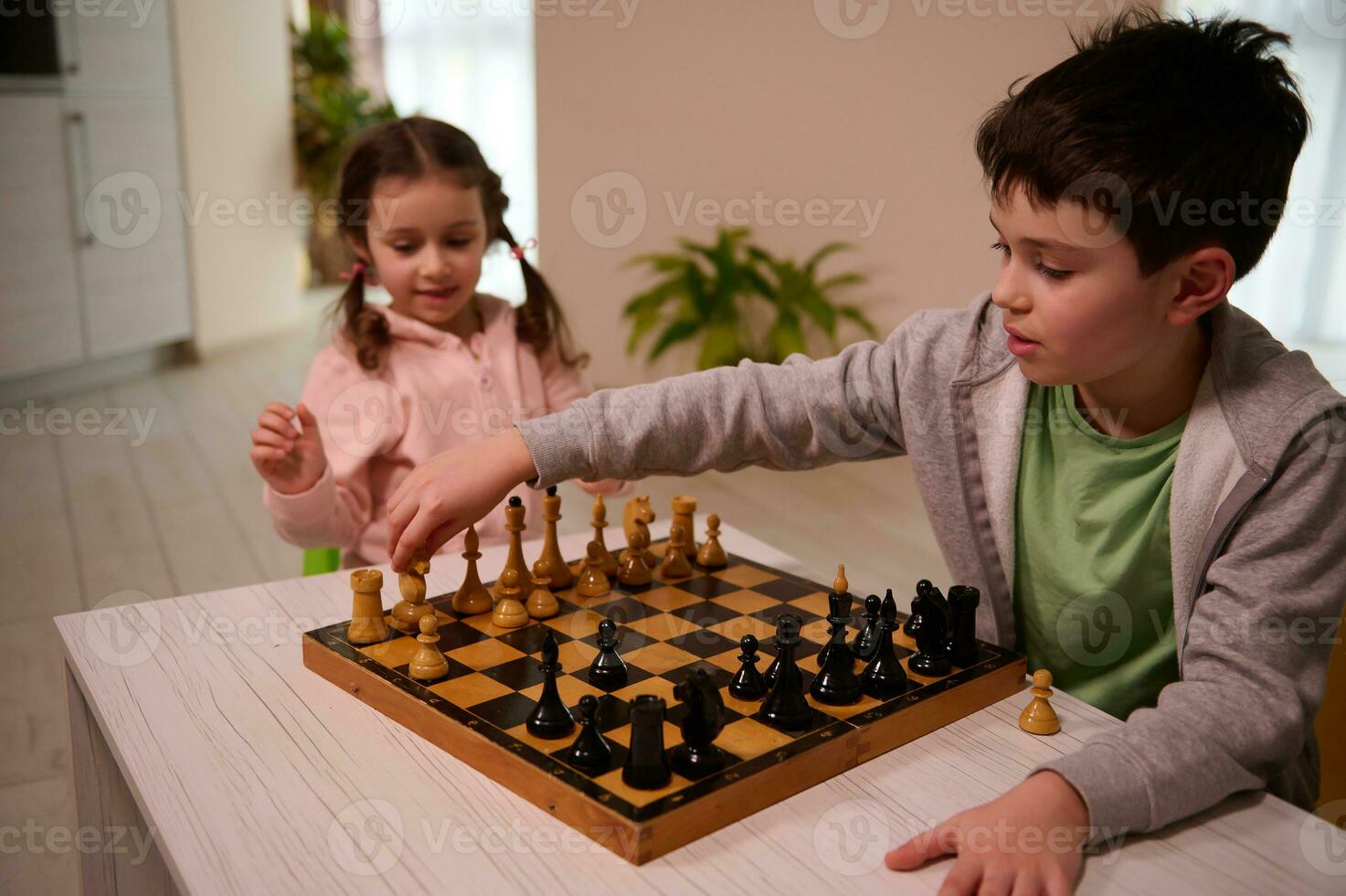 uma fofa Garoto com uma sério Veja senta às a mesa e tocam xadrez com dele mais jovem irmã, colheita acima uma peça do uma cavaleiro e fazer dele mover. cedo desenvolvimento, casa educacional jogos para crianças foto