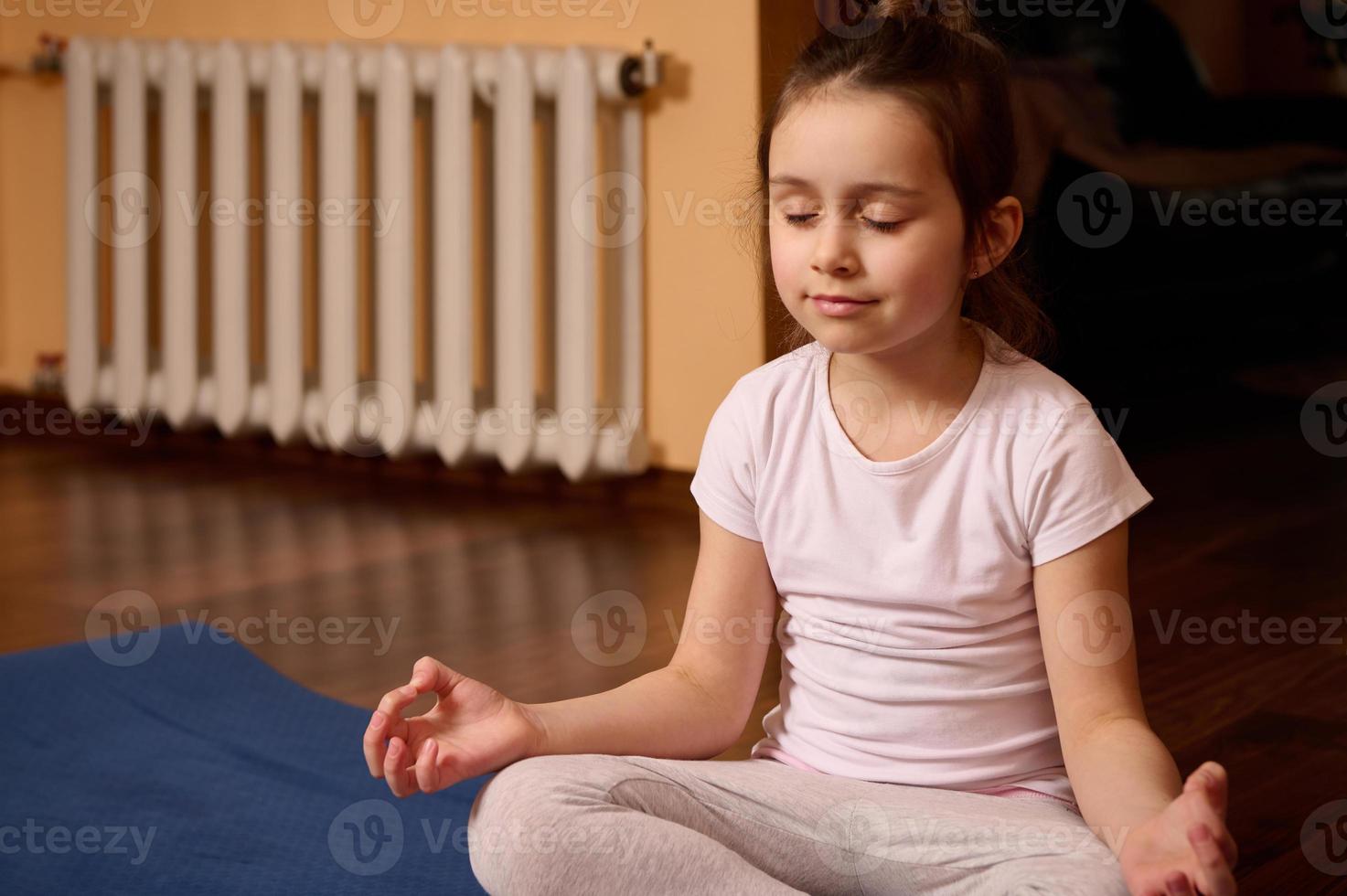 adorável criança menina com fechadas olhos, meditando dentro lótus pose em azul ioga esteira. atenção plena. relaxamento. corpo consciente foto