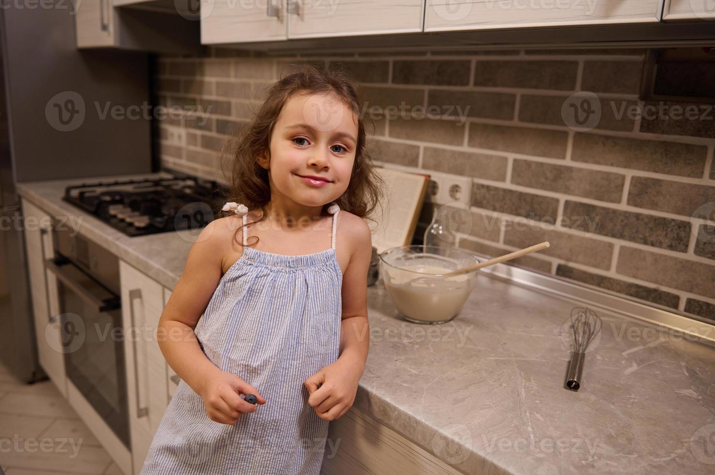 retrato do uma feliz adorável lindo pequeno caucasiano menina dentro azul vestir sorridente fofinho olhando às Câmera, inclinado dela braço em uma cozinha bancada enquanto preparando massa às casa cozinha foto