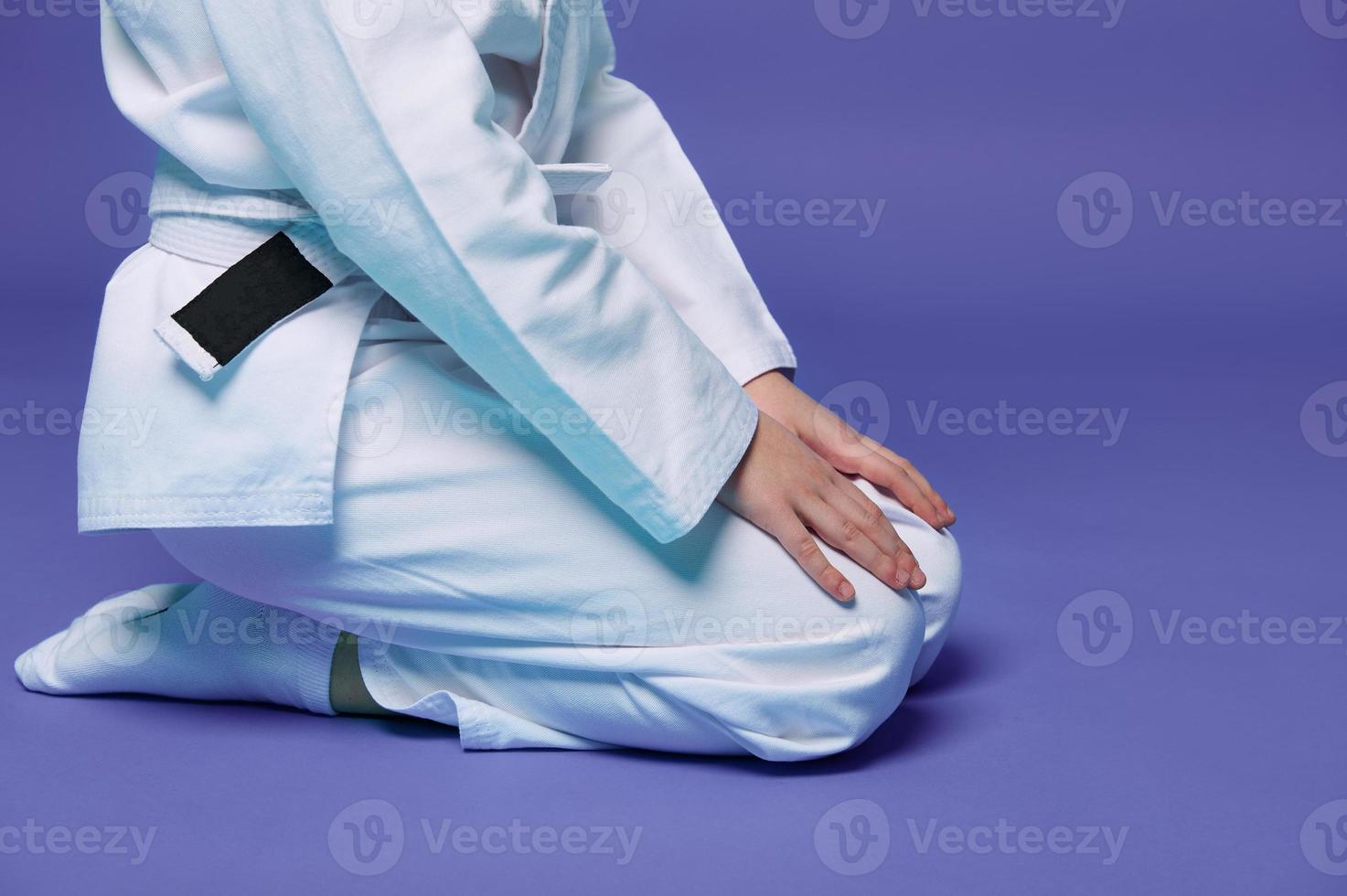 cortada Visão do criança Aikido lutador dentro branco quimono sentado dentro posição com mãos em joelhos isolado em roxa fundo com cópia de espaço. conceito do oriental marcial artes prática foto