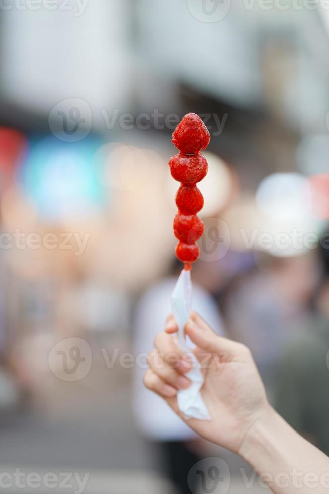 mulher mão segurando caramelo revestido morango espeto às noite mercado. rua Comida e viagem conceito foto