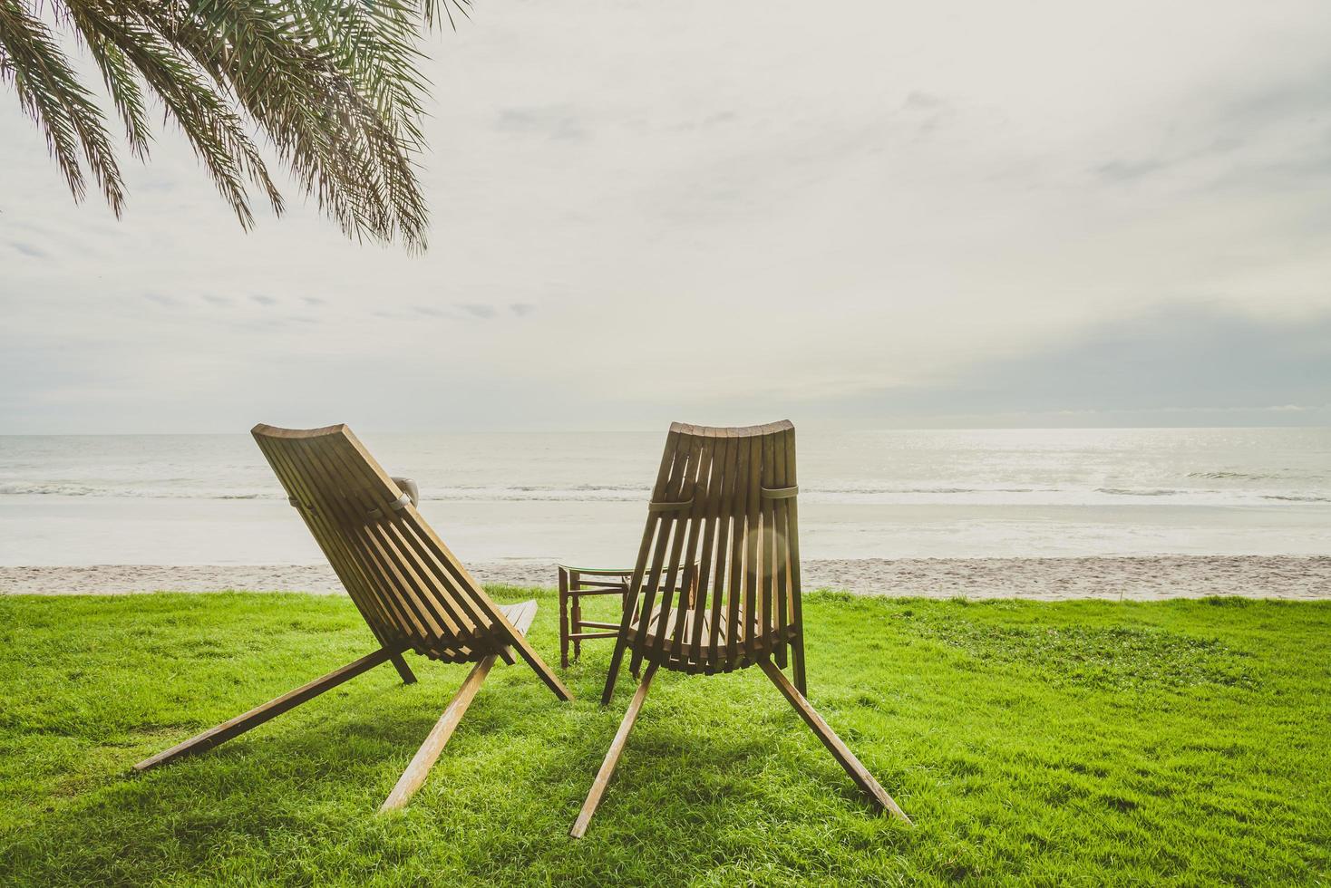 cadeiras de madeira na grama verde com fundo de praia e palmeira foto