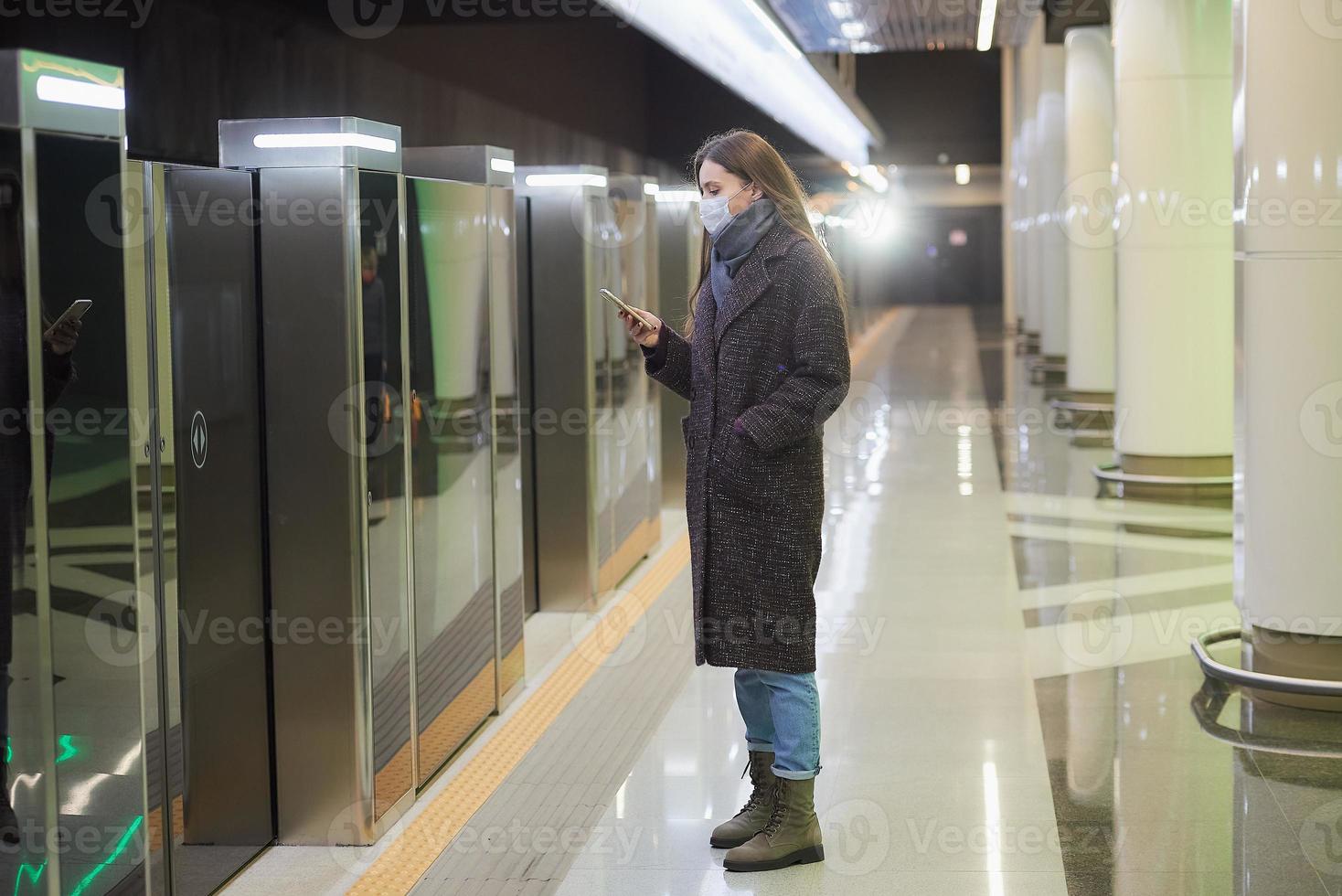 uma mulher com uma máscara facial de médico está esperando a chegada de um trem no metrô foto