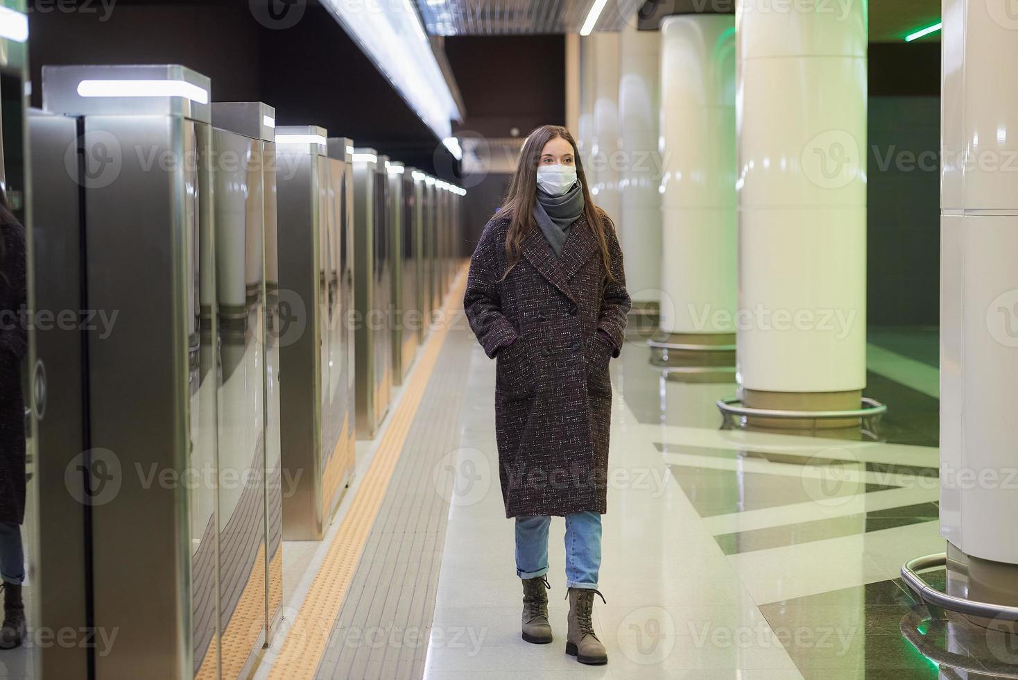 uma mulher com uma máscara facial de médico está esperando a chegada de um trem no metrô foto