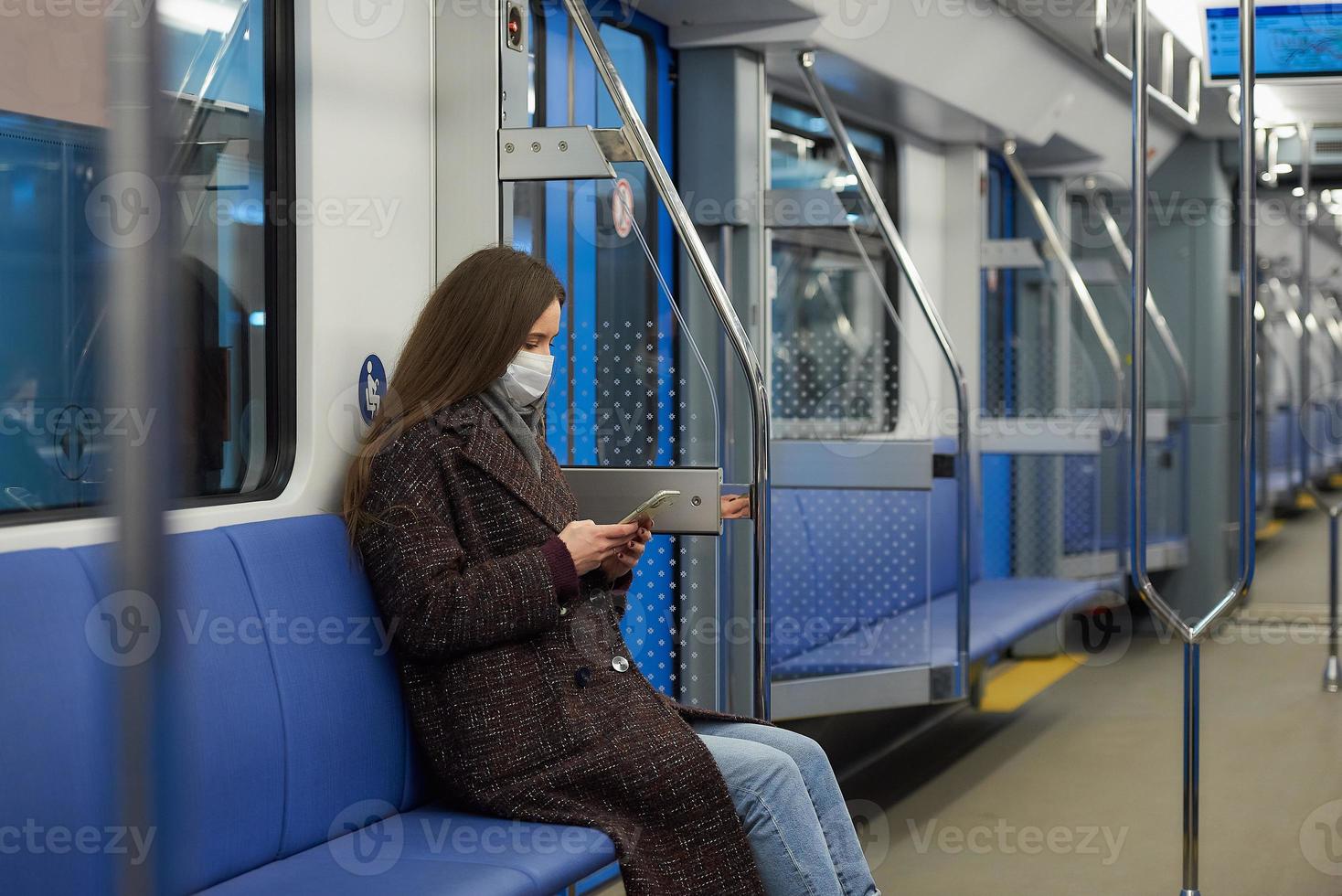 uma mulher com uma máscara facial está sentada e usando um smartphone em um vagão de metrô moderno foto