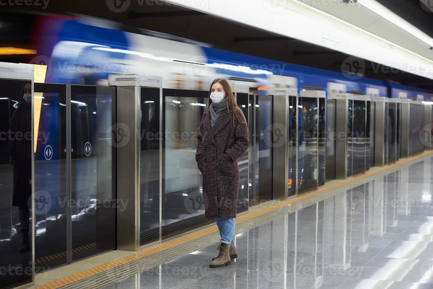 uma mulher com uma máscara facial de médico está esperando a chegada de um trem no metrô foto
