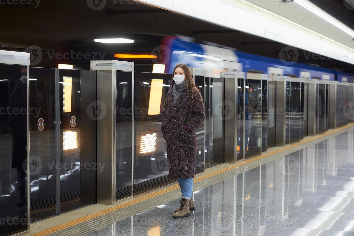 uma mulher com uma máscara facial de médico está esperando a chegada de um trem no metrô foto