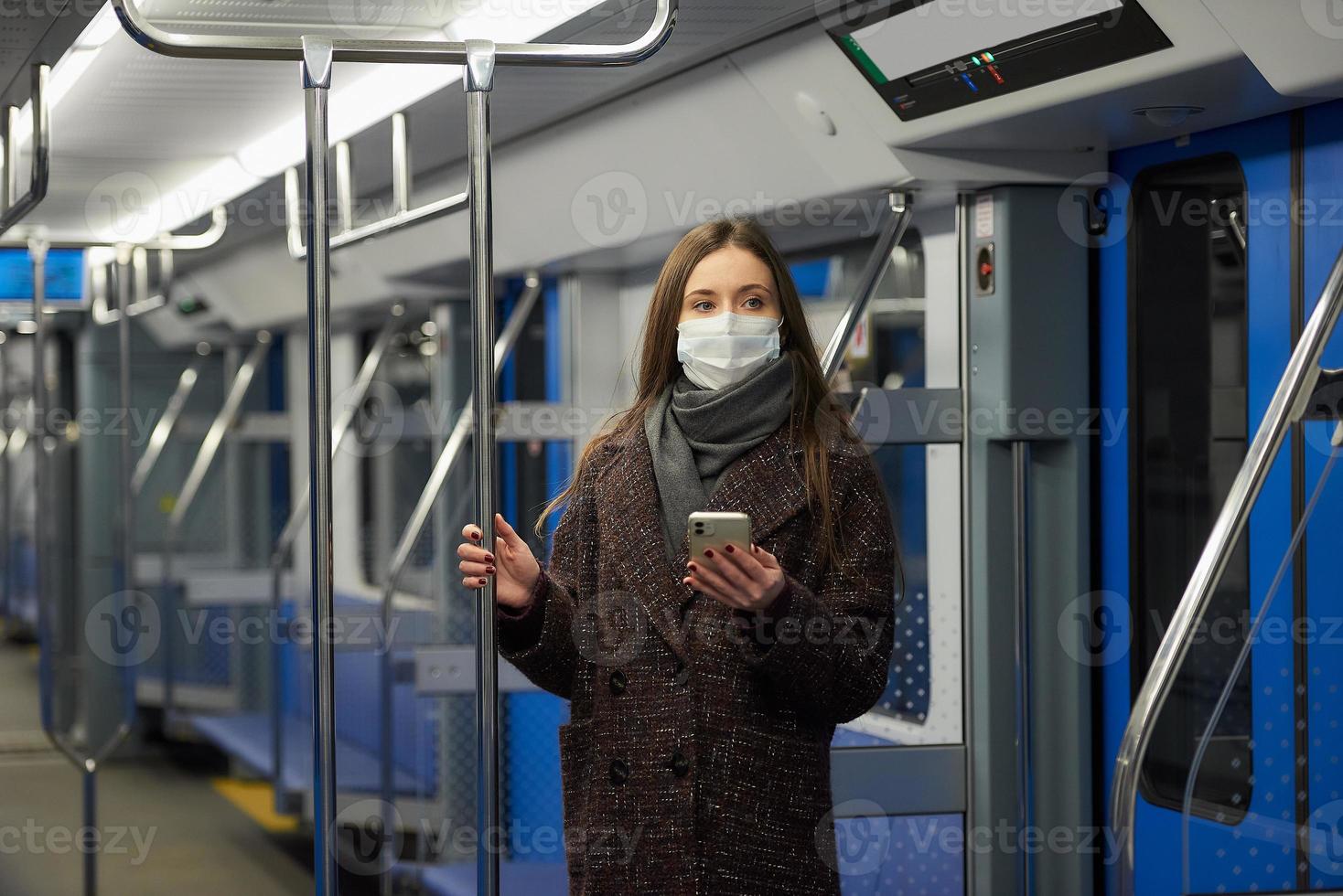uma mulher com uma máscara facial está de pé usando um smartphone em um moderno vagão do metrô foto