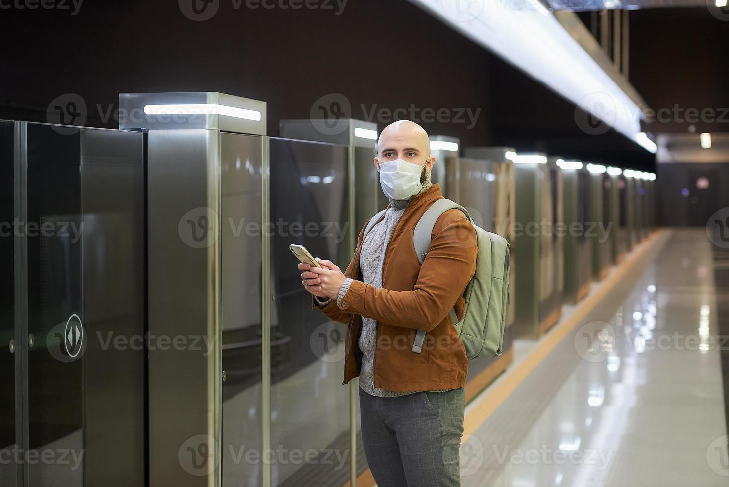 um homem com máscara facial está usando um smartphone enquanto espera o trem do metrô foto