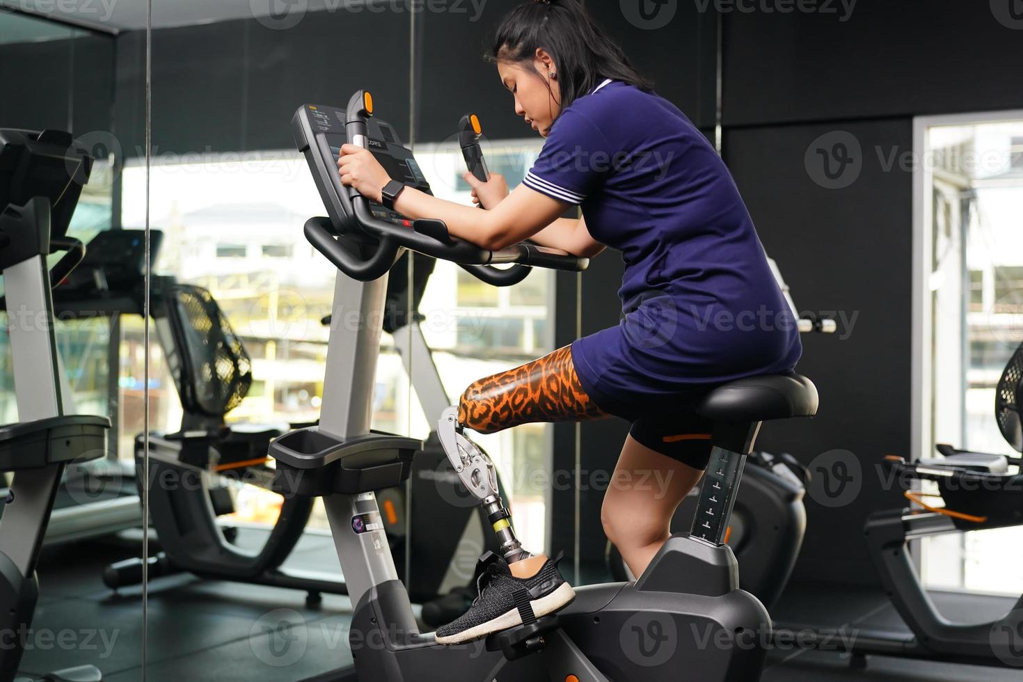 jovem fêmea com 1 protético perna com exercício com uma fiação bicicleta dentro a Academia para prática articulação usar com a de outros normal perna para estar flexível, conceito do vivo uma mulher vida com uma protético foto