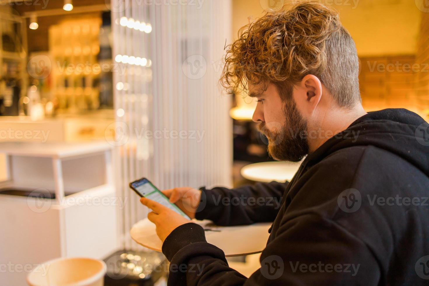 retrato feliz cara fazer dinheiro transação através da inscrição em Móvel telefone com 4g Internet sentado às cafeteria dentro rua noite, sério milenar homem lendo texto mensagem em Smartphone foto