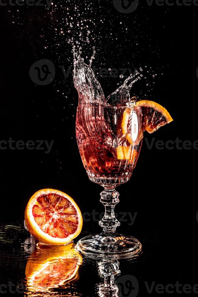 pedaços de laranja cor de sangue caindo em um copo com água espelhada no fundo preto foto