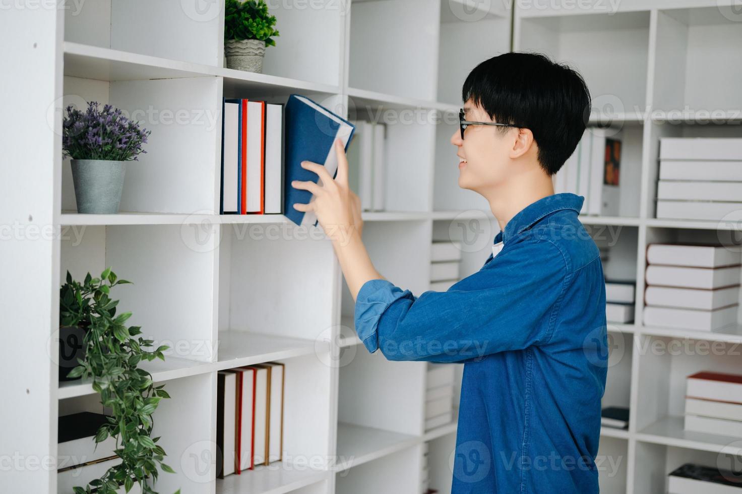 masculino aluna levando notas a partir de uma livro às biblioteca, jovem ásia sentado às escrivaninha fazendo atribuições dentro Faculdade biblioteca foto