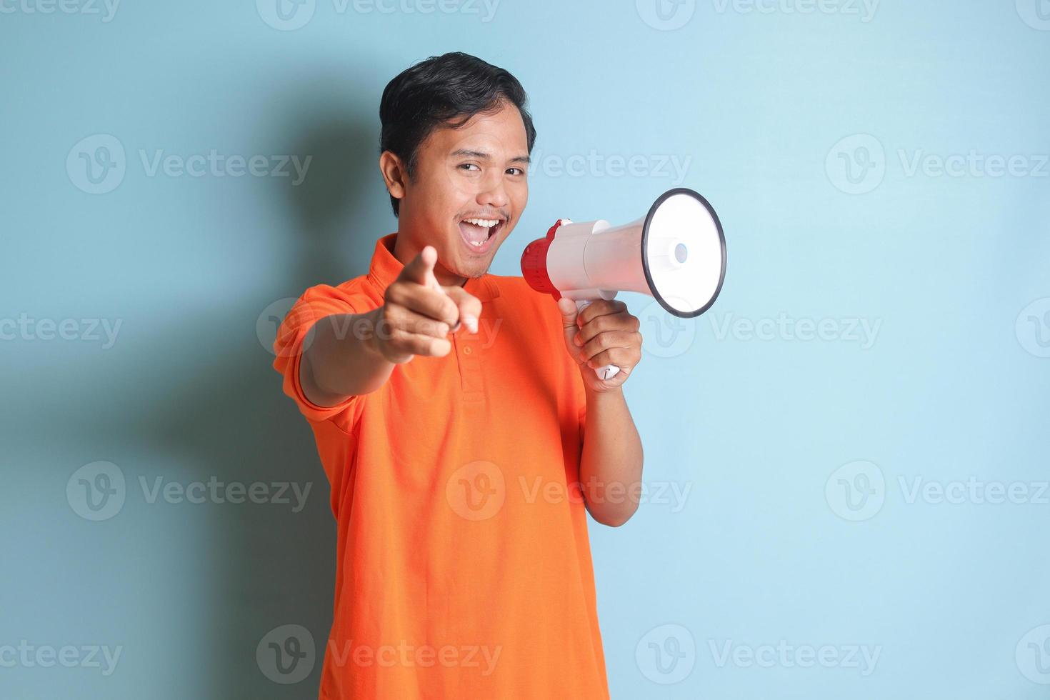 retrato do atraente ásia homem dentro laranja camisa Falando mais alto usando megafone, promovendo produtos. publicidade conceito. isolado imagem em azul fundo foto