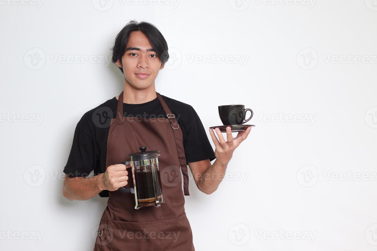 retrato do atraente ásia barista homem dentro Castanho avental mostrando uma copo e pires enquanto segurando francês pressione café criador. isolado imagem em branco fundo foto