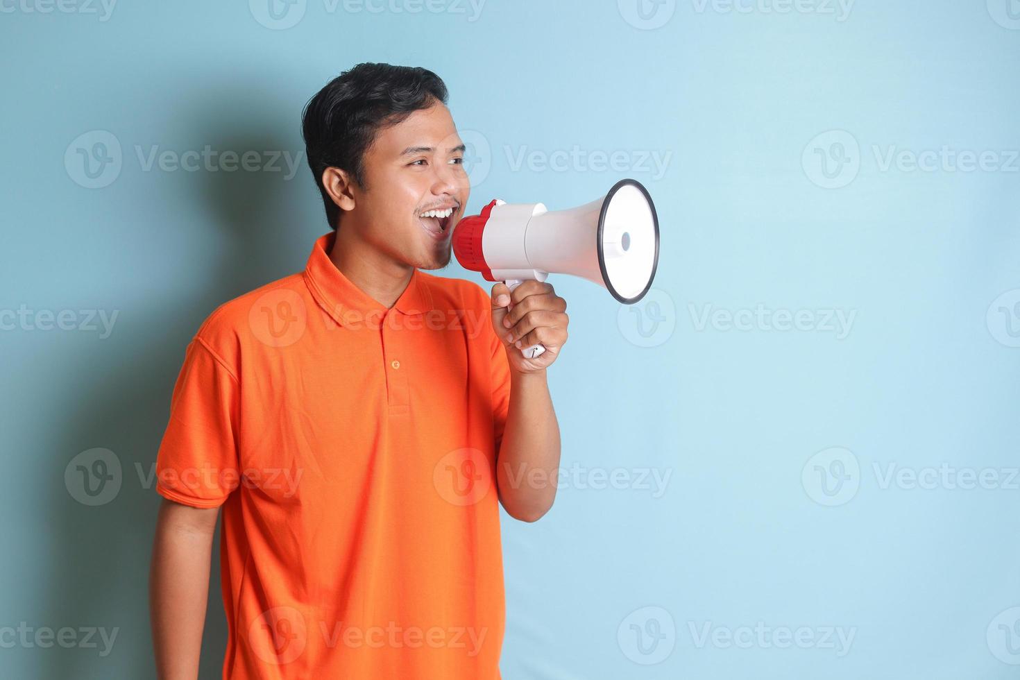 retrato do atraente ásia homem dentro laranja camisa Falando mais alto usando megafone, promovendo produtos. publicidade conceito. isolado imagem em azul fundo foto