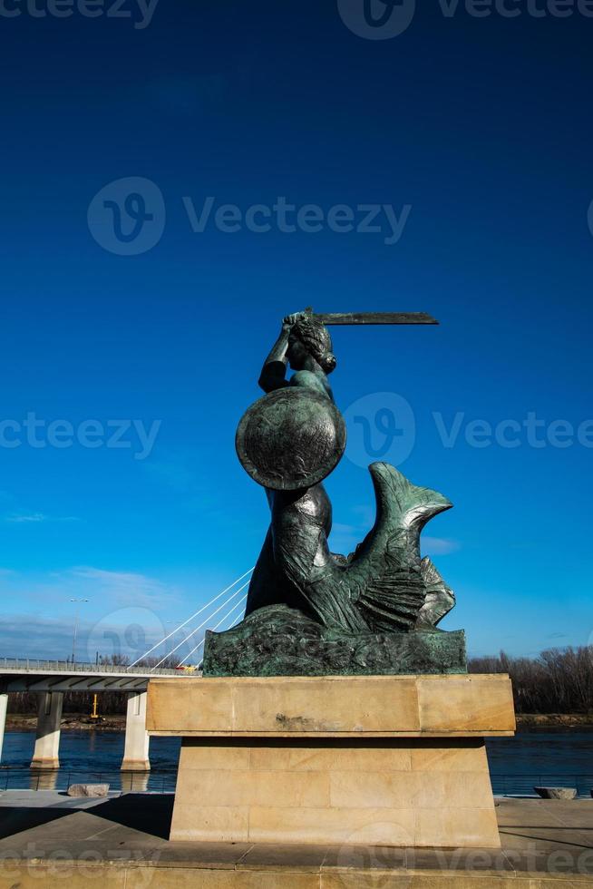 estátua do a Varsóvia sereia em a bancos do a rio com uma ponte dentro a fundo foto