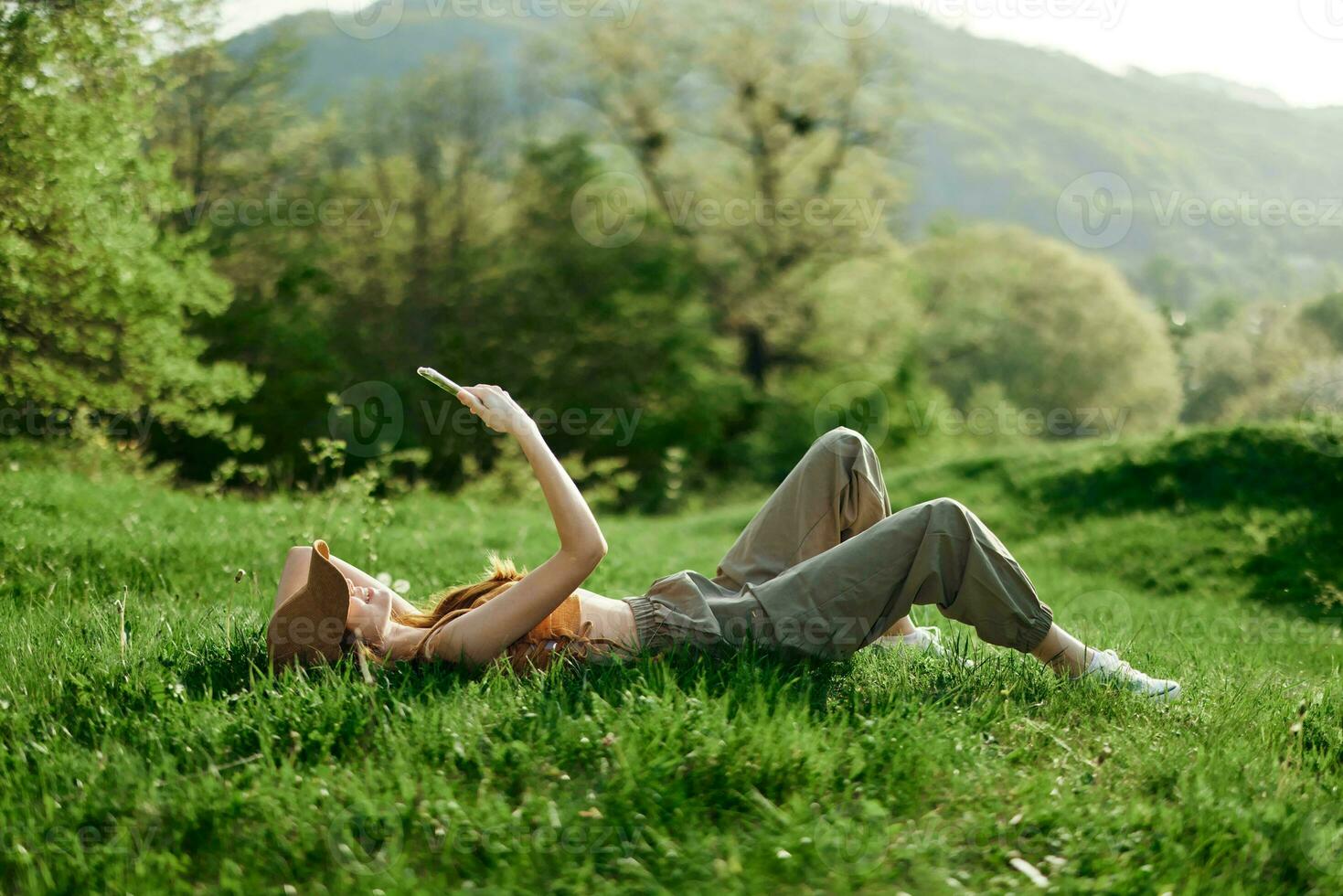 feliz mulher blogueiro deitado em a Relva dentro a parque e sorridente com dela telefone dentro dela mãos contra a pano de fundo do uma verão natural panorama com luz solar foto