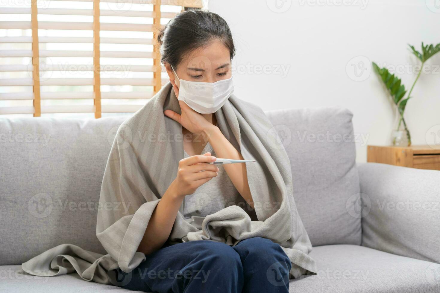 doente, gripe ásia jovem mulher, menina vestindo face mascarar dentro dor de cabeça ter uma febre, gripe e Verifica termômetro a medida corpo temperatura, sentir doença sentado em sofá cama às lar. saúde Cuidado, COVID-19. foto
