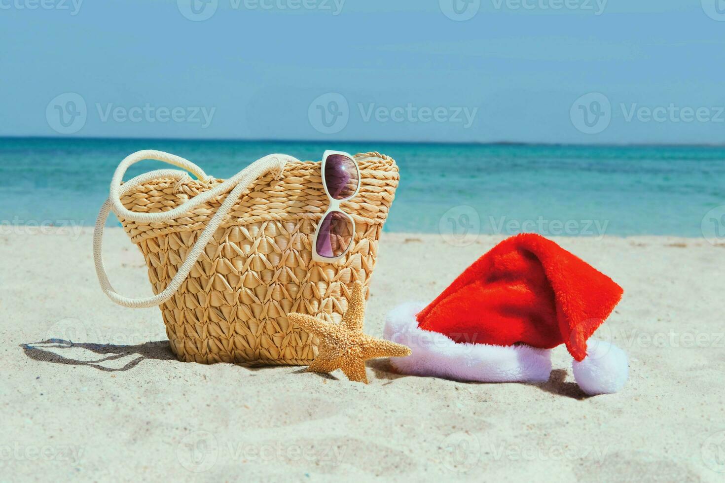 Natal às mar. mulheres de praia acessórios em a areia para período de férias conceito. Palha bolsa, santa claus chapéu, oculos de sol, estrela do Mar. viagem fundo. foto