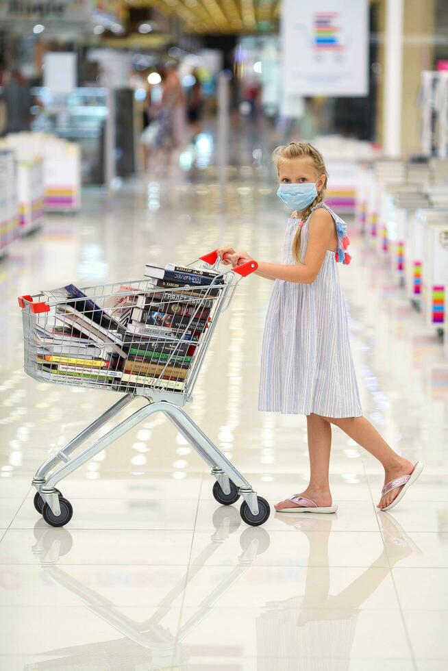 krasnodar, Rússia Julho 16, 2020. a criança dentro uma médico mascarar tem preenchidas uma cesta do livros. pequeno menina dentro a livro loja. oferta. criança dentro a biblioteca com uma carrinho do livros. foto