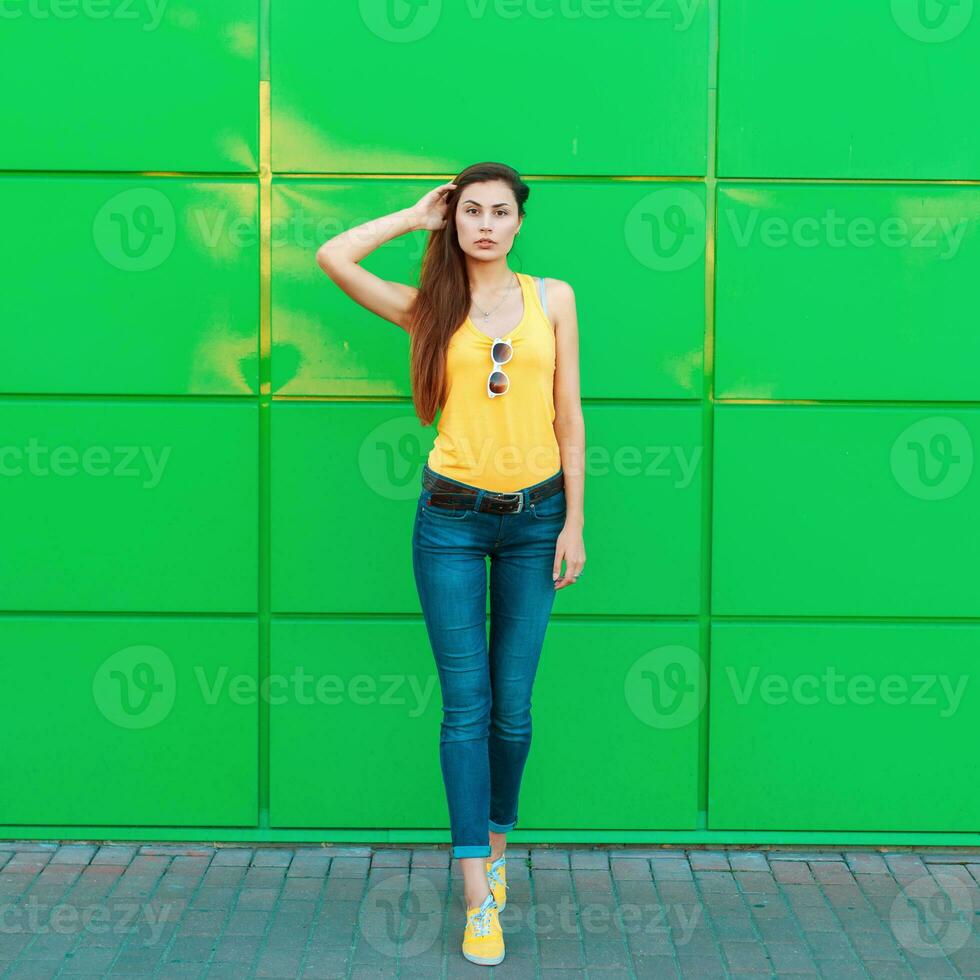 à moda lindo jovem mulher dentro uma amarelo camiseta, jeans e amarelo sapatos posando perto uma brilhante verde parede foto